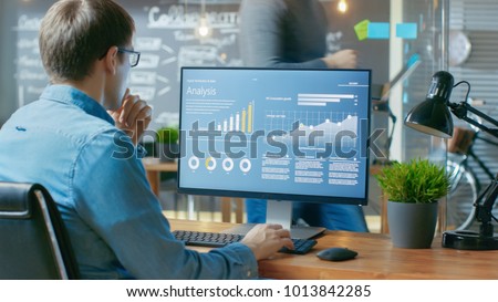 Analyst Works on a Personal Computer Showing Statistics, Graphs and Charts. In the Background His Coworker and Creative Office.
