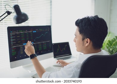 Analyzing data. Back view of young businessman or trader pointing on the data on computer screen with pen while working his modern office.