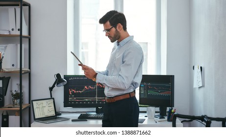 Analyzing data. Successful young trader in formal wear is looking at trading graphs and reports on his digital tablet while standing in front of computer screens with trading charts in the office.