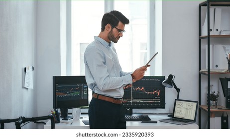 Analyzing data. Successful young trader in formal wear is looking at trading graphs and reports on his digital tablet while standing in front of computer screens with trading charts in the office.