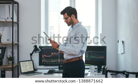 Analyzing data. Successful young trader in formal wear is looking at trading graphs and reports on his digital tablet while standing in front of computer screens with trading charts in the office.