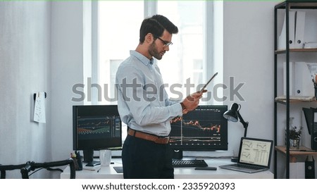 Analyzing data. Successful young trader in formal wear is looking at trading graphs and reports on his digital tablet while standing in front of computer screens with trading charts in the office.