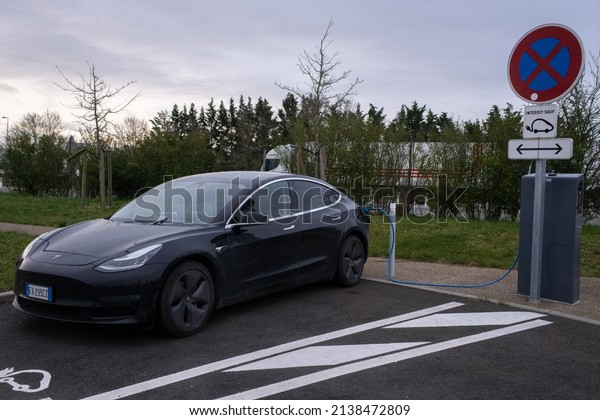 Ancenis,\
France - March 3, 2022: A static shot of a solid black Tesla Model\
3 dual motor long range awd charging at the AC Freshmile charging\
station in cloudy winter day. Selective\
focus.