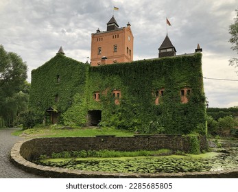 an ancient castle with a tower, overgrown with foliage, on the shore of a lake with lilies, the flag of Ukraine on the tower. Red brick castle. In the foreground is a small lake covered with liliches.