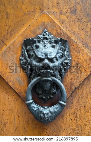 Ancient door knocker in the form of an evil mystical face on a wooden door in Florence, Italy. Ornate Tuscan door knocker in Gothic style. Vintage view of the small old sculpture for knock.