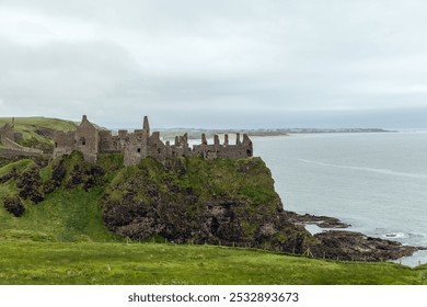 Ancient Dunluce Castle rests on a cliffside by the sea, with rocky outcrops and a coastal village visible in the distance, beneath an overcast sky - Powered by Shutterstock