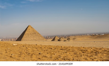 Ancient Egyptian pyramids: a large Menkaura and three small pyramids of queens nearby. In the foreground is the yellow sand of the desert. In the distance- silhouettes of buildings in Cairo. Blue sky.