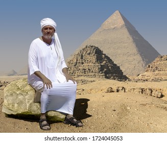 An Ancient Egyptian Sits On A Rock In Front Of The Historic Giza Pyramids In Egypt. Right Behind Him Are Two Little Crumbled Pyramids. The Man Is Wearing A White Robe And A Turban.