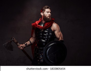 Ancient greek fighter in black armor and red mantle posing standing in dark background and holding his axe and shield.