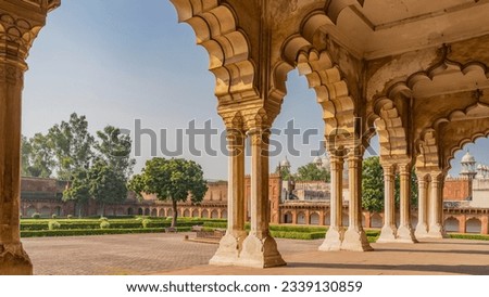 Ancient Indian architecture. Pavilion in the courtyard of the Red Fort. Carved columns with openwork arches against the blue sky. An old cannon is visible on the square. Trimming bushes in the garden