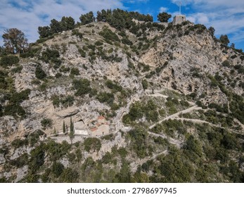 An ancient monastery is nestled into the side of a rugged, rocky mountain cliff, surrounded by lush green vegetation. Winding paths ascend the steep