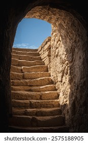An ancient passageway with weathered stone steps ascends into bright daylight. The texture of the rough stones contrasts with the clear blue sky, hinting at history and exploration.