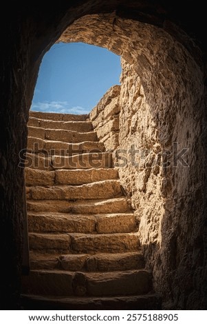 An ancient passageway with weathered stone steps ascends into bright daylight. The texture of the rough stones contrasts with the clear blue sky, hinting at history and exploration.