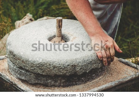 The ancient quern stone hand mill with grain. The man grinds the grain into flour with the help of a millstone. Men's hands on a millstone. Old grinding stones turned by hands