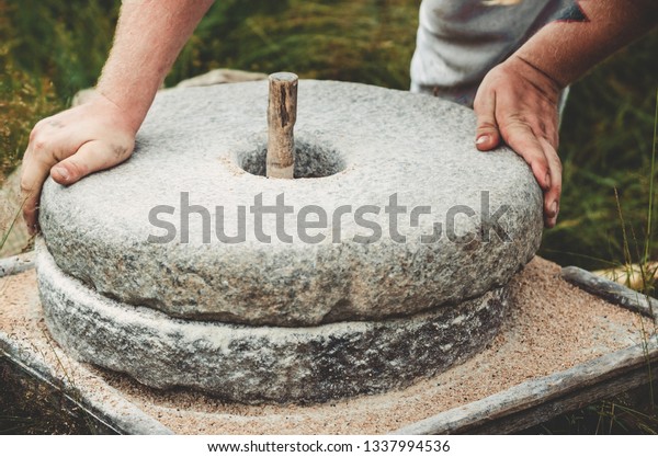 The\
ancient quern stone hand mill with grain. The man grinds the grain\
into flour with the help of a millstone. Men\'s hands on a\
millstone. Old grinding stones turned by\
hands