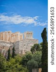 Ancient ruins of the Acropolis in Athens, Greece, stand majestically against a bright blue sky with scattered clouds, framed by lush green trees