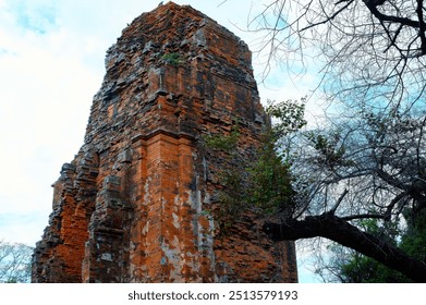 Ancient Ruins, History, Decay, Nature, Resilience, Time, Architecture, Angkor, captures a weathered, ancient brick tower set against a bright sky, with a stark, leafless tree framing its side - Powered by Shutterstock