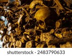 An ancient skull, bathed in shadows and long dead and decayed, sits atop a pile of old bones in the dark depths of the Catacombs of Paris, France.