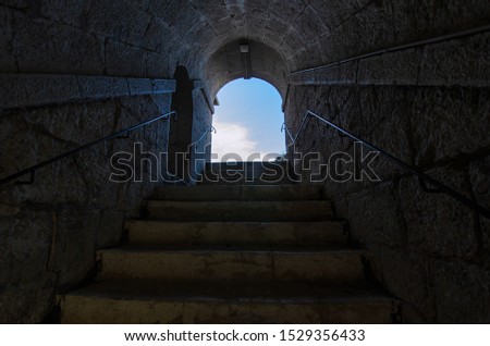 An ancient staircase inside the tunnel leads up. There are stone steps. The walls of the tunnel are made of stone blocks. At the end of the stairs you can see the blue sky. Background.