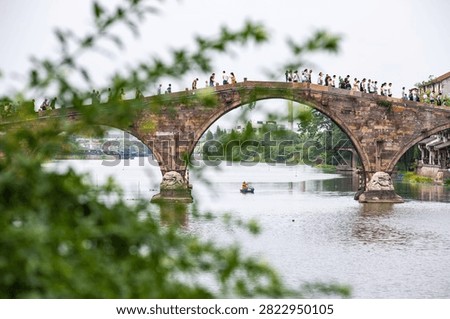 Ancient stone arch bridge spanning calm river with tourists walking across, framed by green foliage and traditional riverside architecture in historic Chinese town.