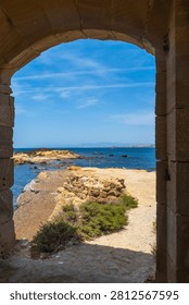 Ancient stone archway frames the rugged coastline of tabarca island, revealing the deep blue mediterranean sea under a clear summer sky, offering a glimpse of the historic protected marine reserve