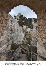 Ancient stone archway framing a dramatic descent of modern metal staircases through the layered ruins of a medieval hilltop castle, with cypress trees and a lone tower rising against an overcast sky