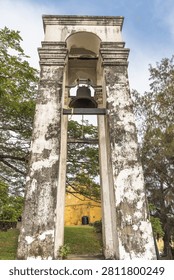 Ancient stone bell tower with a large metal bell against a clear sky, surrounded by green trees historic architecture. Galle, the historic port city on the southwest coast of Sri Lanka.