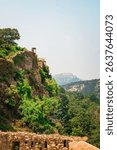 Ancient stone fort walls covered in natural greenery, overlooking the forested valley and mountains, depicting historical architecture blending with Indian nature