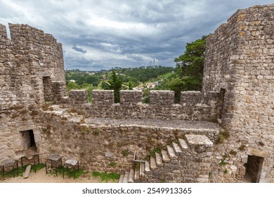 ancient stone fortress wall with battlements and staircases, surrounded by lush greenery and hills under a dramatic cloudy sky, evoking a sense of history and resilience - Powered by Shutterstock