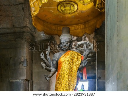 Ancient stone statue of multi-armed Hindu deity draped in gold cloth inside Angkor Wat temple. Altar with offerings, incense and golden ceremonial umbrella. Religion and spirituality concept.