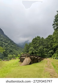 Ancient stone structure surrounded by lush green grass, dense forest, and mountain valley landscape under a misty cloudy sky. Peaceful outdoor nature scenery with historical ruins and natural environment.