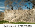 Ancient stone wall featuring two sets of weathered stairs, leading upwards under a clear blue sky and several bare trees, conveying a sense of history and ascent