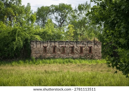 Ancient stone wall with vertical loop holes standing on grassy meadow surrounded by green trees inside historical Lednice castle park scenic landscape view