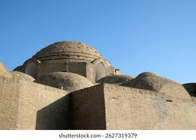 An ancient structure features multiple domes and solid stone walls under a bright blue sky. The architecture reflects historical significance and cultural heritage of the region.