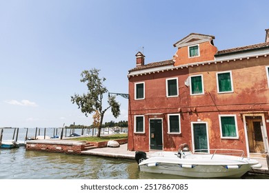 an ancient terracotta-colored building with green shutters on the bank of a canal, next to a moored boat. The backdrop features clear blue skies, calm water and green trees, creating the atmosphere of - Powered by Shutterstock