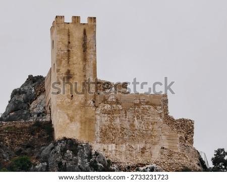 Ancient tower keep rises from rocky base, weathered stone blocks and battlement lines revealing medieval