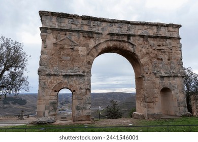 ancient, weathered stone arch with two openings stands against a backdrop of a cloudy sky and distant landscape - Powered by Shutterstock