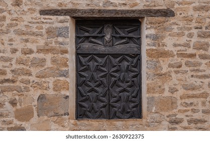 An ancient wooden window set in a weathered stone wall, located in a historic village in Spain. The detailed geometric patterns of the woodwork contrast beautifully with the rough texture of the stone