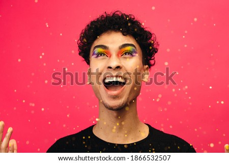 Androgynous male with rainbow eye makeup throwing up the glitter and laughing. Excited gay man with glitters flying against red background.