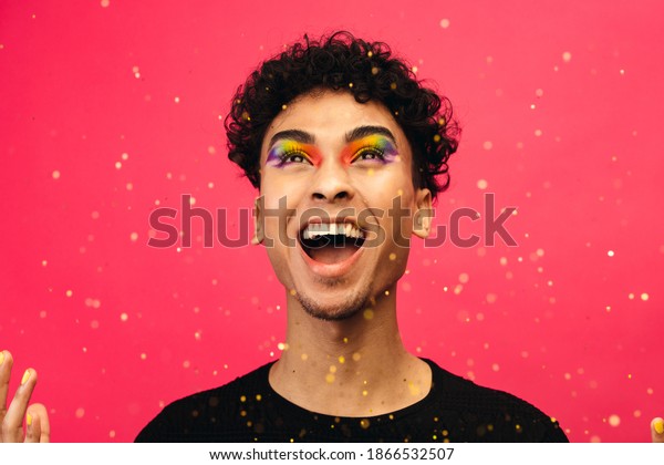 Androgynous male with rainbow eye makeup\
throwing up the glitter and laughing. Excited gay man with glitters\
flying against red\
background.