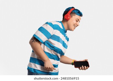 An androgynous person with blue hair and red headphones, wearing a blue and white striped shirt, holding a smartphone and smiling, studio setting, representing joy, music, and LGBT pride. - Powered by Shutterstock