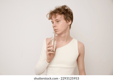 Androgynous young red haired man in white designer crop top holds a glass bottle with bamboo tube in hand drinks water supports ecology and looks away while standing against gray background in studio. - Powered by Shutterstock