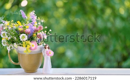 angel figurine and flowers bouquet in mug on table outdoor, green natural background. Religious church holiday. symbol of faith in God, christianity. Happy Name Day greeting card design. copy space