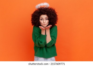 Angelic woman with Afro hairstyle wearing green casual style sweater and nimbus over head sending air kisses, expressing love and romance. Indoor studio shot isolated on orange background.