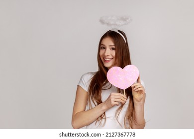 Angelic woman with holy nimbus holding pink paper heart on stick and smiling to camera, demonstrating love care, romantic feelings, wears white T-shirt. Indoor studio shot isolated on gray background.