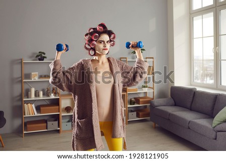 Anger is best motivator. Funny angry woman having sports workout at home. Determined housewife in homewear, curlers and beauty skincare face mask doing fitness exercise with dumbbells in living-room