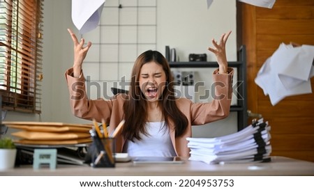 Angered, furious, crazy and mad millennial Asian businesswoman or female office worker screaming, shouting and throwing up papers at her office desk. Depression, overworked, failure.