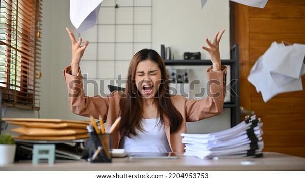 Angered,\
furious, crazy and mad millennial Asian businesswoman or female\
office worker screaming, shouting and throwing up papers at her\
office desk. Depression, overworked,\
failure.