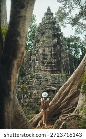 Angkor Thom backpacker in white hat stands among giant tree roots, gazing at a Bayon face tower in lush jungle ruins. Concept, high quality image for travel ads, blogs, social posts and tourism guides
