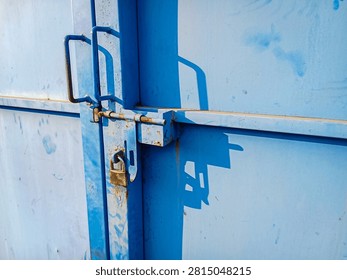 Angle close-up view of a locked padlock on a weather-worn blue iron door with hard shadows cast on the metallic surface.